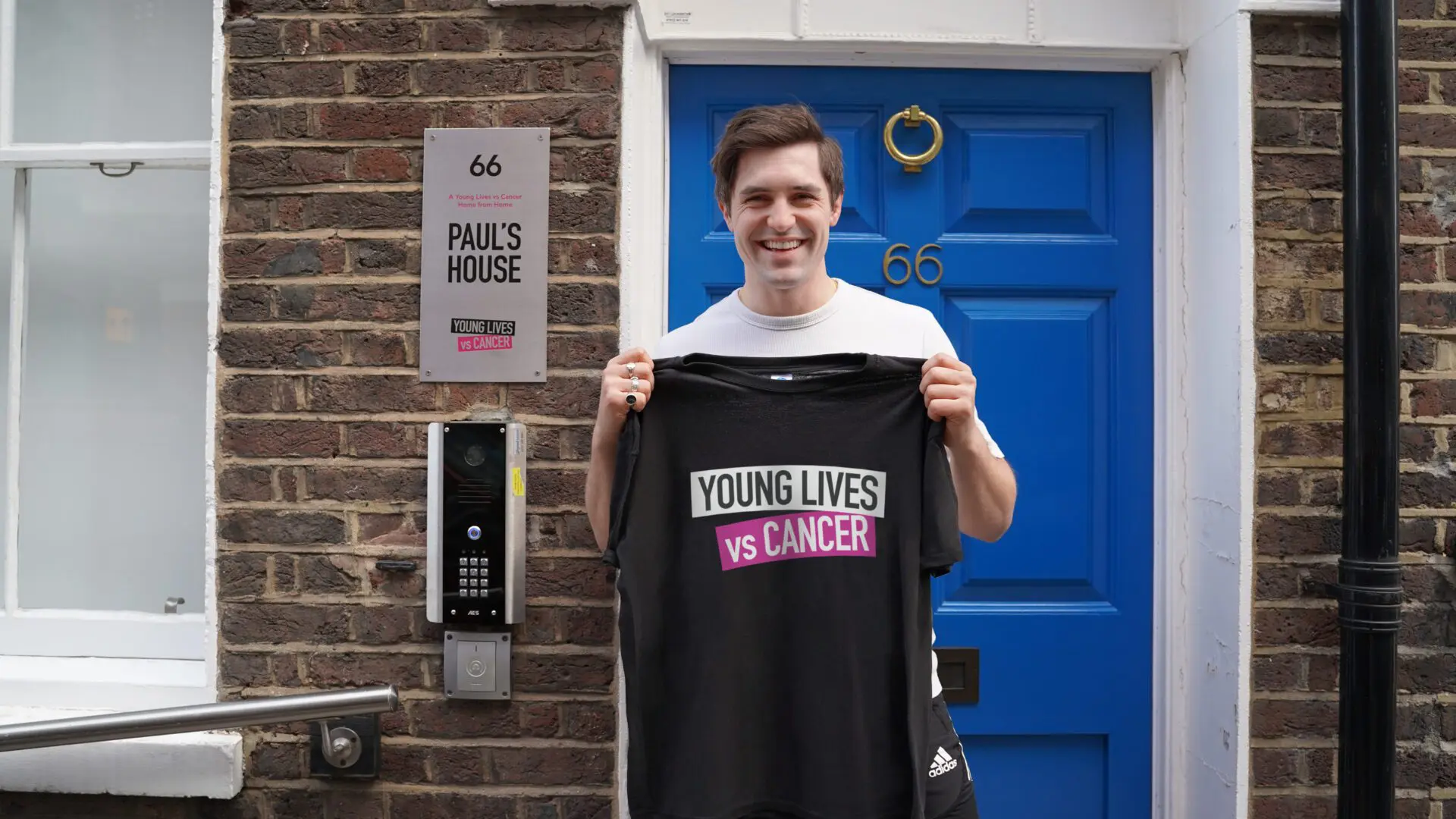 An image of actor Phil Dunster holding a Young Lives vs Cancer t-shirt outside Paul's House, one of Young Lives vs Cancer's Homes from Home.