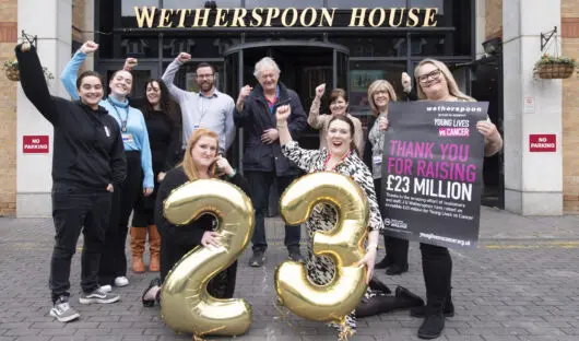 A group of JDW colleagues and Young Lives vs Cancer staff are stood outside Wetherspoon House. Two people are crouched at the front in front of a line of people at the back, they are holding gold 2 and 3 balloons - everyone has their hands up in the air to celebrate.