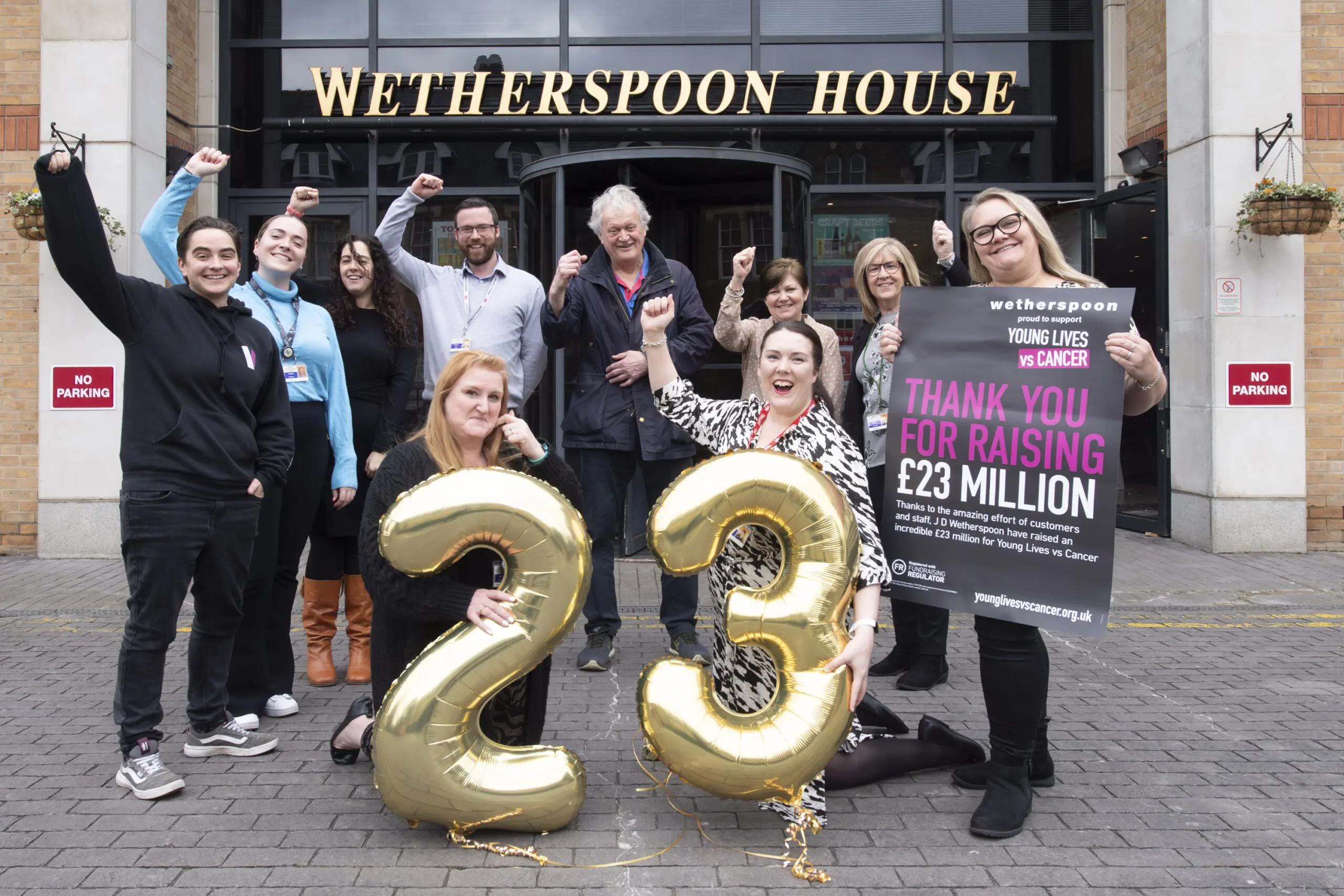 A group of JDW colleagues and Young Lives vs Cancer staff are stood outside Wetherspoon House. Two people are crouched at the front in front of a line of people at the back, they are holding gold 2 and 3 balloons - everyone has their hands up in the air to celebrate.