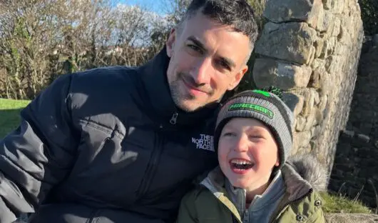 John and his son, Alfie, are sitting on a big rock. Alfie is laughing, while John has his left arm around him. Behind them there is a stone wall.
