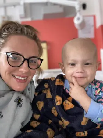 Orla, a young toddler with her Mum Naomi. They are next to each other smiling at the camera.