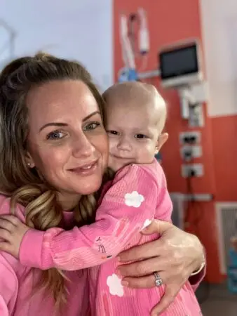 Orla, a young toddler with her Mum Naomi. They are next to each other smiling at the camera.