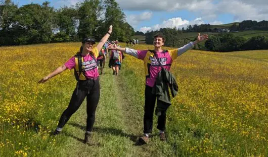 Two people with open arms and big smiles wearing Young Lives vs Cancer T-shirts in a field in summer
