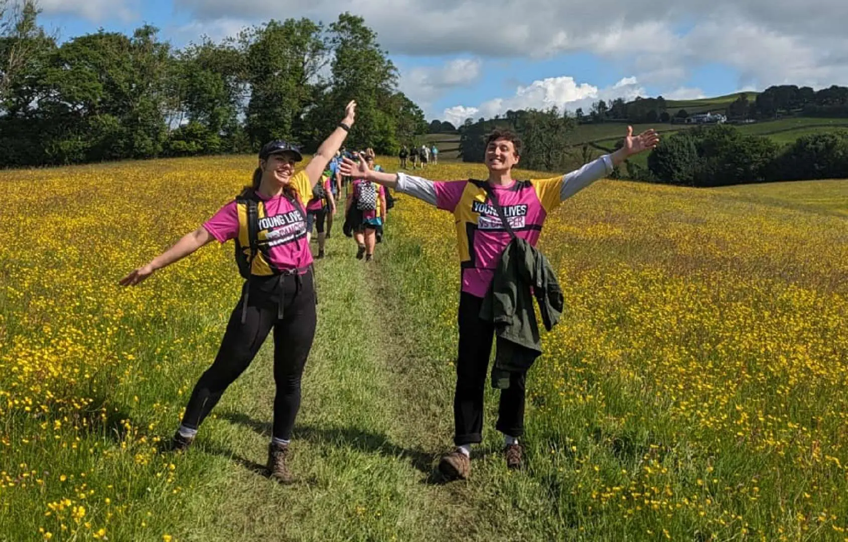 Two people with open arms and big smiles wearing Young Lives vs Cancer T-shirts in a field in summer