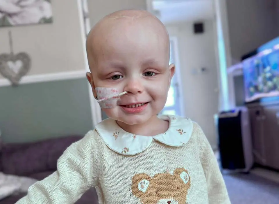 Little girl, Nancy, with no hair smiling at the camera. She is going through treatment for brain cancer and wearing a feeding tube