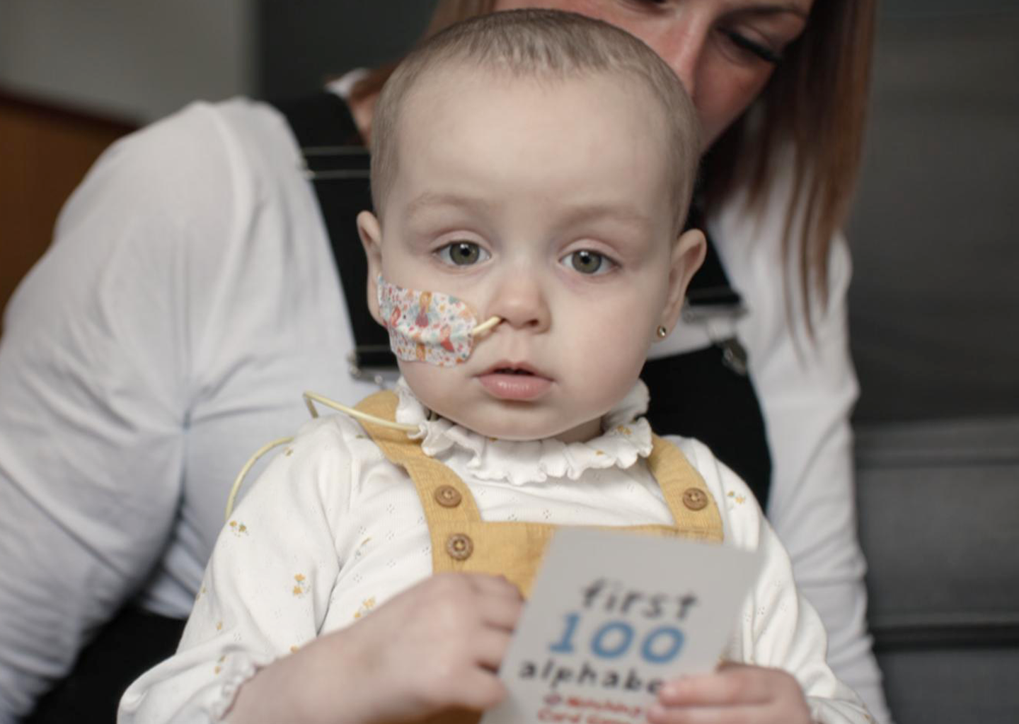 Little girl, Nancy, with no hair looking at the camera sitting on her mothers knee. She is going through cancer treatment and wearing a feeding tube.