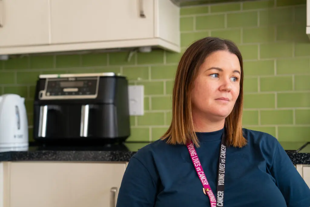 A young lives vs cancer social worker sat in a kitchen looking off to the side. They are wearing a Young Lives vs cancer lanyard.