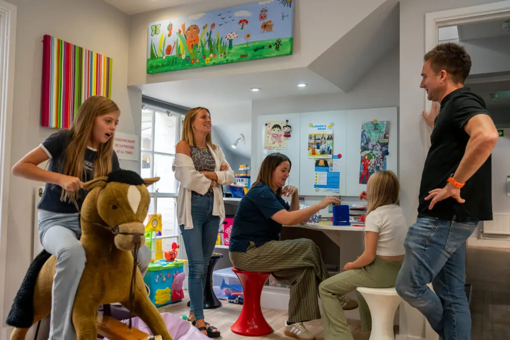 two young people and three adults standing in a playroom. One of the young people is sat at the table playing a boardgame with one of the adults and another is on a rocking horse. 