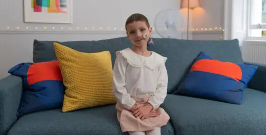 Child sitting on a teal sofa with yellow and blue cushions, wearing a white blouse and pink skirt.