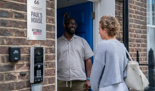A young person standing on the doorstep of Paul's House, a Young Lives vs Cancer home from home accommodation. Paul's House manager is standing welcoming them through the door.