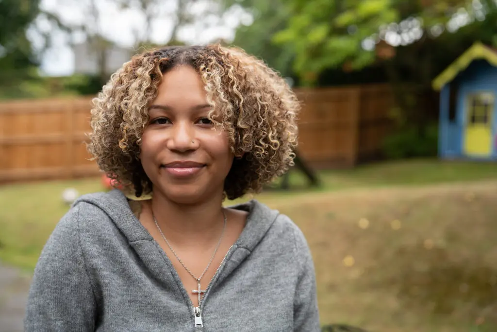 A young person in a grey hoodie stood in a garden smiling at the camera.