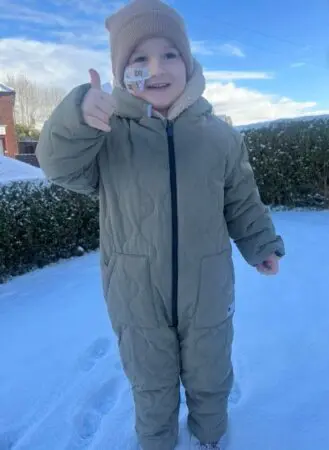 Daithi smiling giving a thumbs up to the camera standing in the snow. He is wearing a snowsuit and beanie hat.