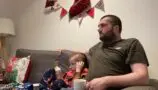 Daithi and his dad sat on a sofa with christmas decorations on the wall above them. They are at Pauls house, a Young Lives vs Cancer Home from Home accommodation.