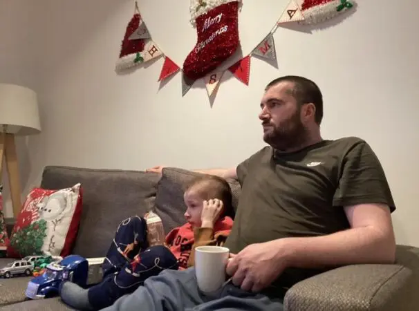 Daithi and his dad sat on a sofa with christmas decorations on the wall above them. They are at Pauls house, a Young Lives vs Cancer Home from Home accommodation.