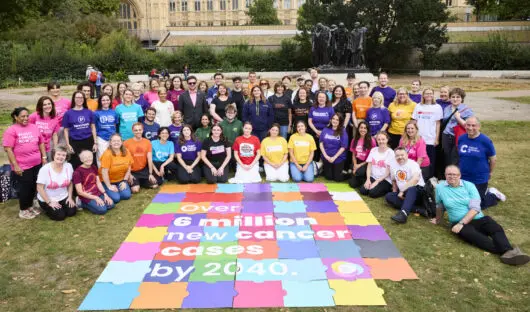 A group of people wearing different coloured cancer charity t-shirts sit around a huge coloured puzzle that says 