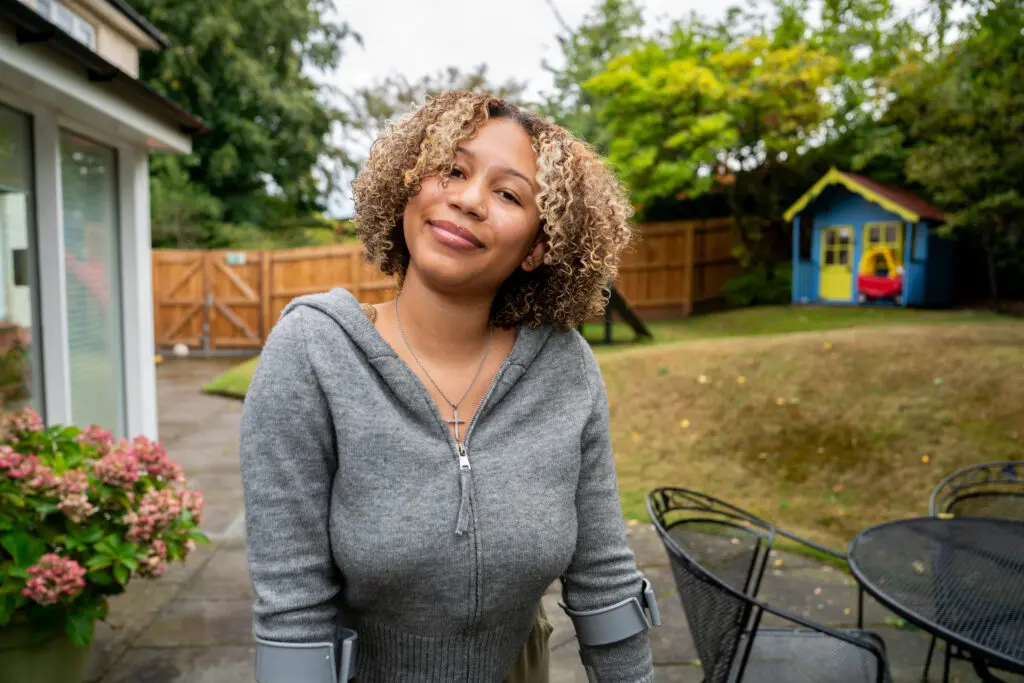 Young person stood in a garden smiling at the camera. They are wearing a grey hoodie and are standing with crutches. 