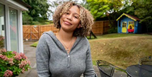 Young person stood in a garden smiling at the camera. They are wearing a grey hoodie and are standing with crutches.