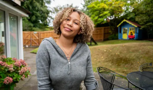Young person stood in a garden smiling at the camera. They are wearing a grey hoodie and are standing with crutches.