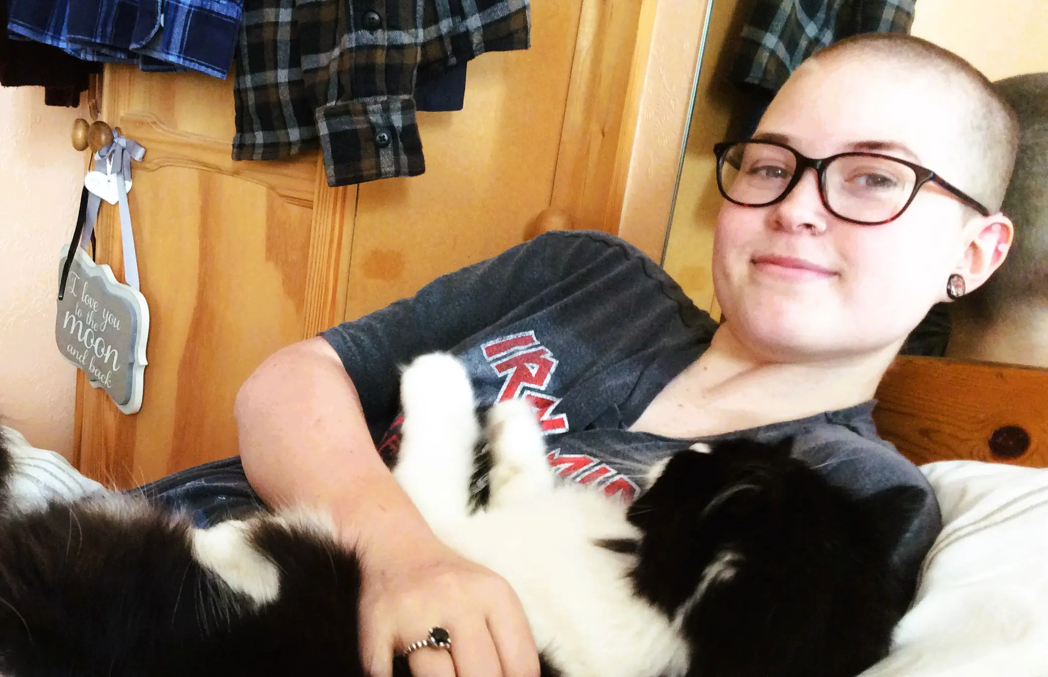 Megan lying on her bed cuddling a black and white cat. She has lost her hair and is wearing glasses, smiling.