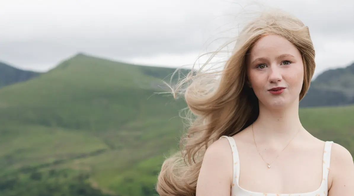 Young person Enlli standing with the Welsh countryside behind her looking towards the camera.