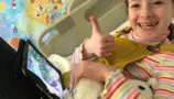 Lucy on the day of her diagnosis lying in a hopsital bed holding a tablet and soft toy giving thumbs up.