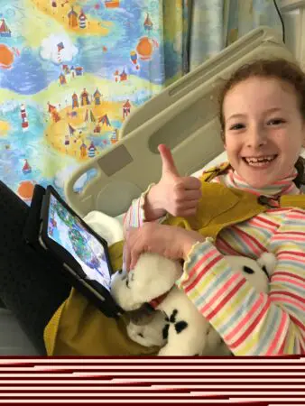 Lucy on the day of her diagnosis lying in a hopsital bed holding a tablet and soft toy giving thumbs up.