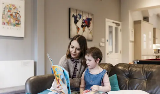 Margot and her mum Georgia sat on a sofa. Georgia is reading to Margot.