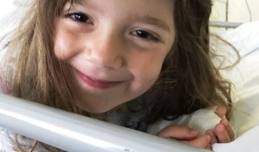 A young girl with cancer peeks through the bars of her hospital bed smiling