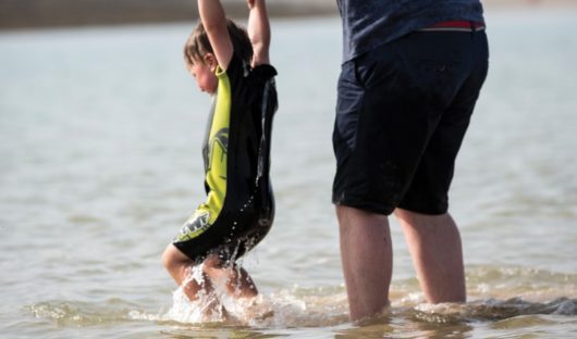 A child with cancer holds his dad's hands as he splashes in the sea
