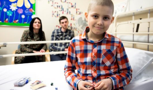 A boy with cancer sits on the edge of his hospital bed smiling. His parents are sitting in the background.