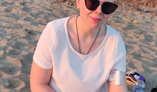 Young cancer patient Vic sitting cross legged on the beach in sunglasses