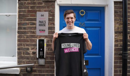 An image of actor Phil Dunster holding a Young Lives vs Cancer t-shirt outside Paul's House, one of Young Lives vs Cancer's Homes from Home.