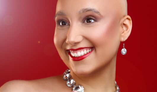 Amy is standing against a red backdrop, she has her head shaved, make up on and a large necklace and earrings