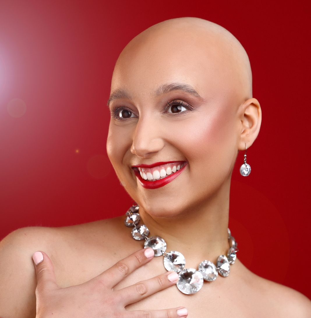 Amy is standing against a red backdrop, she has her head shaved, make up on and a large necklace and earrings