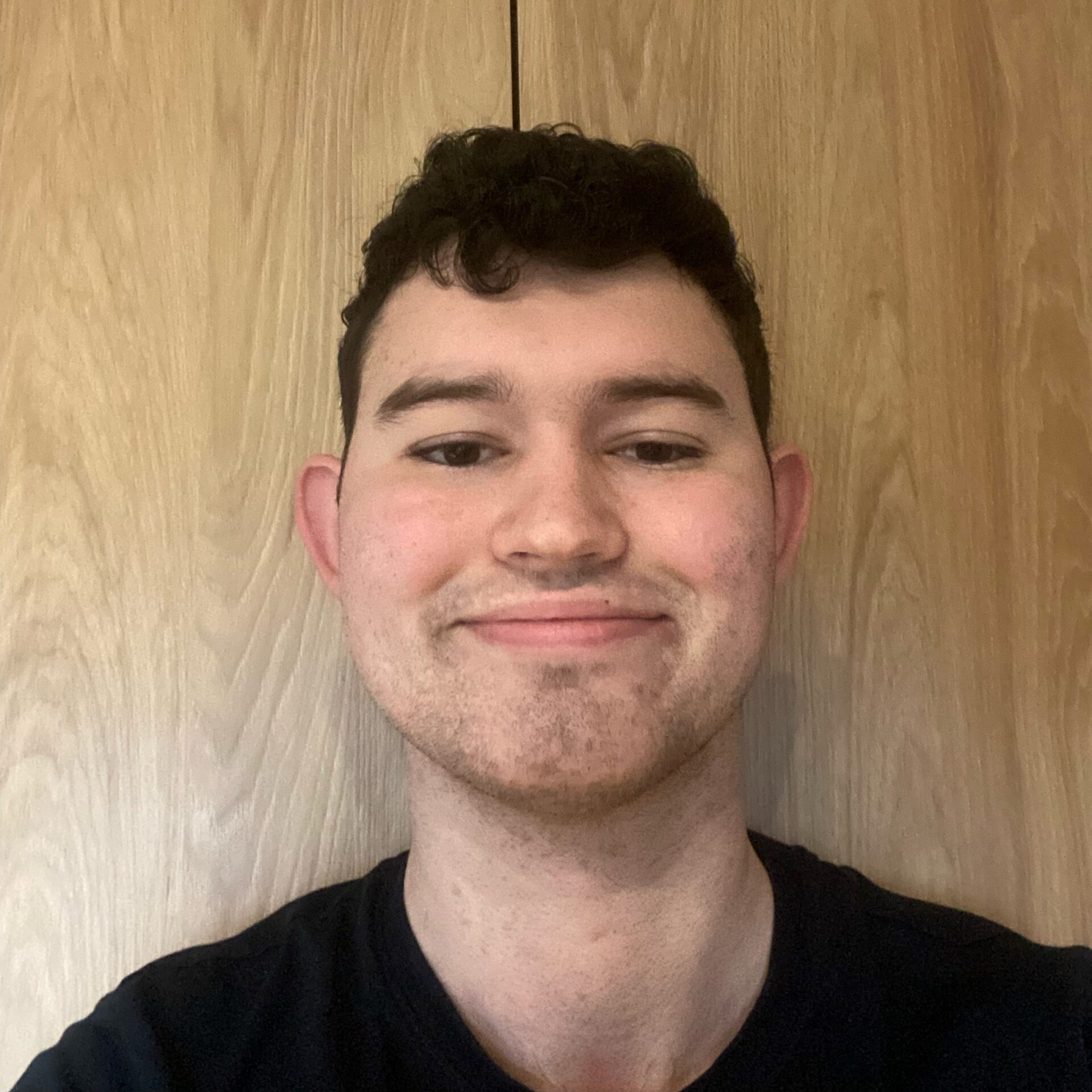 Quinn is standing infront of a wooden background, he is wearing a black t-shirt and smiling at the camera