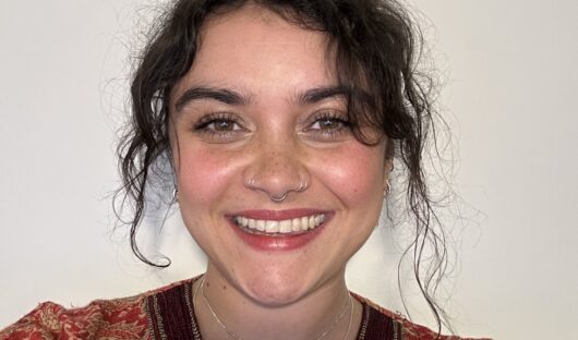 This is a selfie featuring a woman, Emma. She has curly brown hair and is wearing an orange top with yellow and brown patterns.